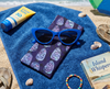 Canvas glasses case featuring a semi abstract design based on bubbleweed in purples and blues on a small blue towel on a beach with a pair of blue sunglasses, a book and sunblock