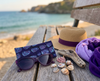 Canvas glasses case featuring a semi abstract design based on bubbleweed in purples and blues on a wooden bench by the sea, along with a pair of purple glsses, a sun hat, purple scarf, driftwood and shells