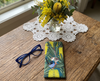 Canvas glasses case featuring a stylised pair of superb fairywrens in a Banksia marginata on a wooden bench with a pair of blue glasses and a vase of yellow native Australian flowers
