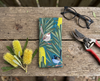 Canvas glasses case featuring a stylised pair of superb fairywrens in a Banksia marginata on a wooden bench with a pair of glasses, a pair of secateurs and some cut banksia flowers