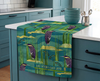 linen tea towel covered with stylised print of black bitterns and black catfish against a watery background with reed and rocks lying n a bench in a blue kitchen