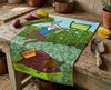 linen tea towel printed with a comic style picture of a man shaking a rake at an orange-footed scrub fowl that is digging up his garden on a bench with vegetables and a garden trowel