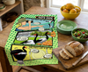 linen tea towel printed with a comic book style layout showing magpie geese eating mangos or being eaten themselves on a kitchen table with a bowl of mangos, a salad and a loaf of bread