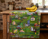Linen tea towel with an all-over pattern of tropical houses in a lush landscape on a bench with a bunch of bananas