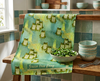 a linen teatowel featuring stylised green tree frogs on a watery green background with some small clumps of grass in a kitchen scene