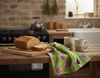 In a rustic kitchen, a linen tea towel with a pattern of comb-crested jacanas, sacred lotus flowers and lilypads lies between two steaming mugs and a cut loaf of banana bread on a chopping board
