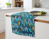 a linen tea towel printed with a pattern of colourful lures against blue and green waves on a kitchen bench with a plate of fish