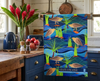 linen tea towel featuring a stylised pattern of Nankeen night herons in a blue stream with rocks, reeds and pandanus hanging in a rustic kitchen