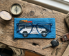 Canvas glasses slip case featuring a troop carrier with a loaded roof rack on a blue mottled background. Sitting on a timber bench beneath a pair of blue glasses with a compass and a set of car keys