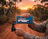 Canvas glasses slip case featuring a troop carrier with a loaded roof rack on a blue mottled background, held up in a man's hand along with a pair of dark glasses against a dirt track in the Australian bush at sunset