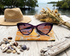 A canvas glasses slip case showing a stylised male and female white-breasted whistler on a mangrove flat sits on a timber tabletop under a pair of purple sunglasses surrounded by driftwood, a bunch of dried Australian native flowers, shells and driftwood. In the background is a mangrove scene at high tide