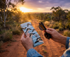 Canvas glasses slip case featuring several pop art bush stone curlews with a speech bubble saying 'Well, what?' against a pale blue striped background, held in a man's hand against a sunset in an Australian bush scene with a track. A second hand holds a pair of sunglasses. 