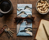 Canvas glasses slip case featuring several pop art bush stone curlews with a speech bubble saying 'Well, what?' against a pale blue striped background. A pair of brown sunglasses sits on top of the glasses case which is surrounded by a notebook, a bowl of unshelled peanuts, a bunch of keys and a mug of black coffee