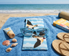 Canvas glasses slip case featuring two stylised welcome swallows in flight against a simple cloud pattern in pale blue with a pair of sunglasses on a towel on a beach with a book, some sandals, sunblock and shells