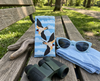 Canvas glasses slip case featuring two stylised welcome swallows in flight against a simple cloud pattern in pale blue propped up on a piece of driftwood on a bench in a park with sunglasses, a scarf and a pair of binoculars