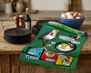 linen tea towel printed with a comic book style layout describing plovers attacking people in parks to protect their chicks lying on a bench in a kitchen with plates and a bowl of eggs